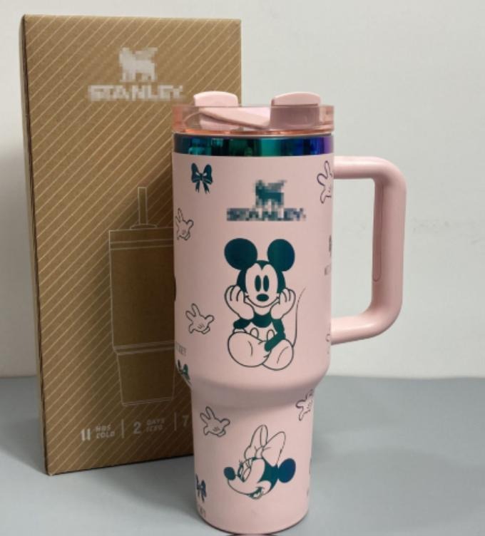 Gourde Stanley Flowstate Tumbler 40 oz avec anse — rose pâle avec motifs bleu-vert et bande métallique irisée, motif personnage rond assis à grande tête et